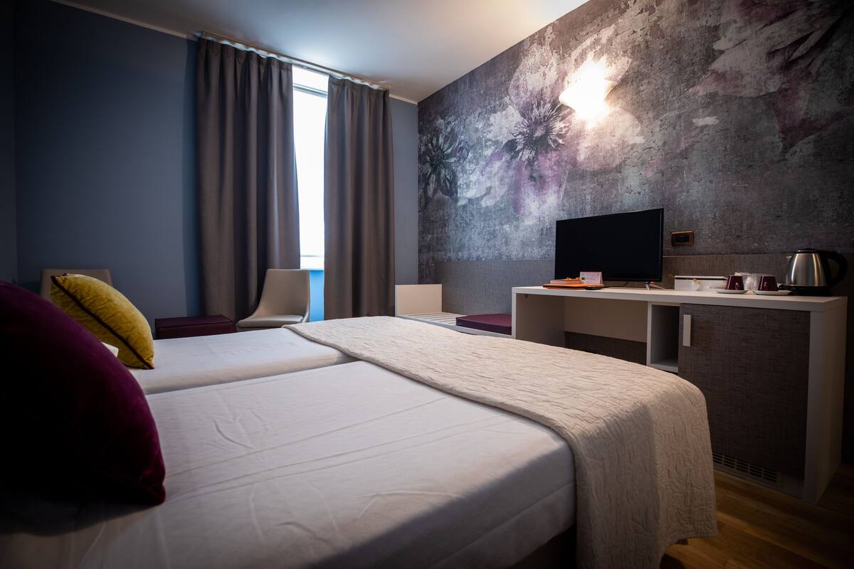 A modern hotel room with two beds, a desk, a flat-screen TV, and a large abstract mural on the wall.