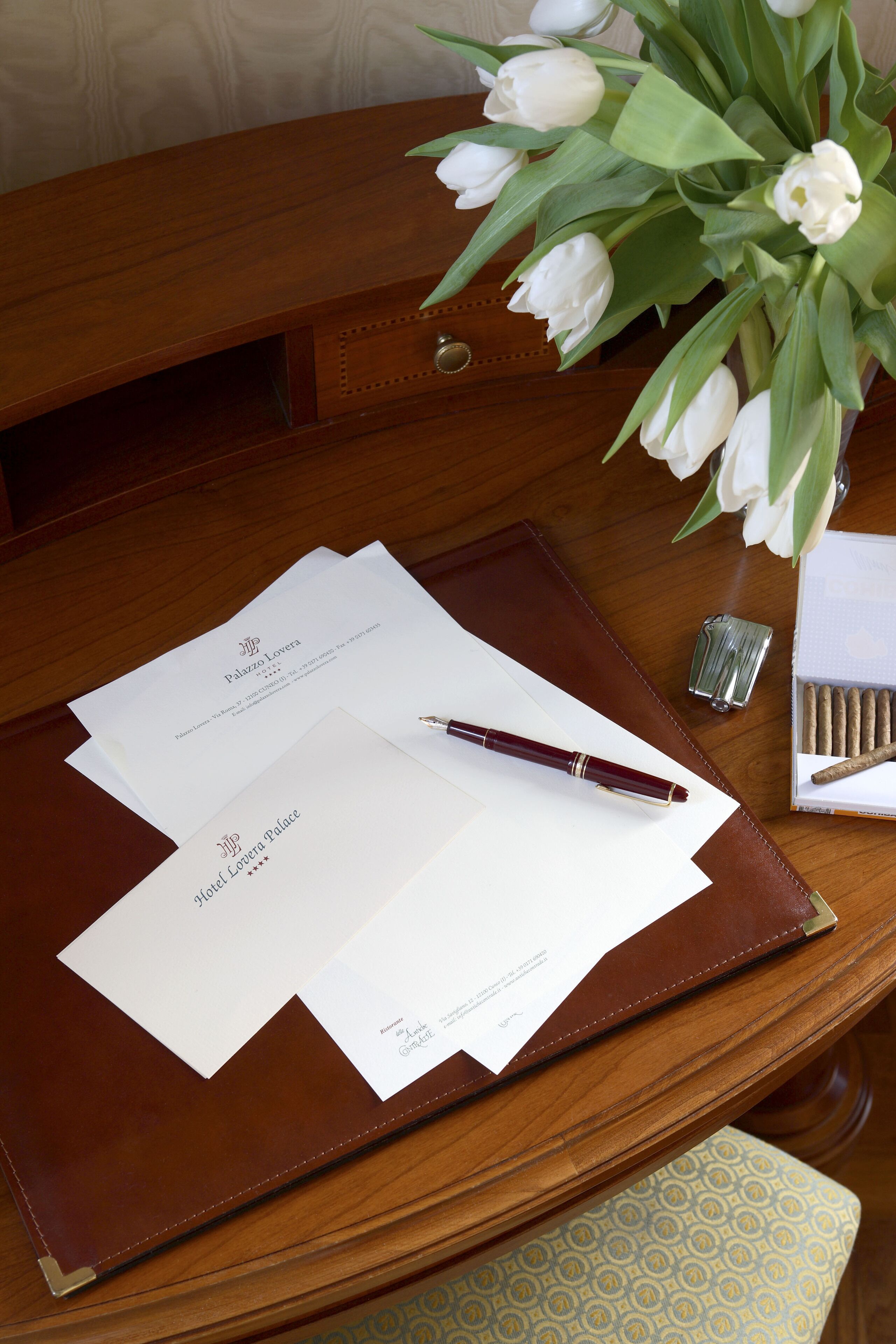 A wooden desk with a brown leather folder, two white papers, a pen, a lighter, and a box of cigarettes.