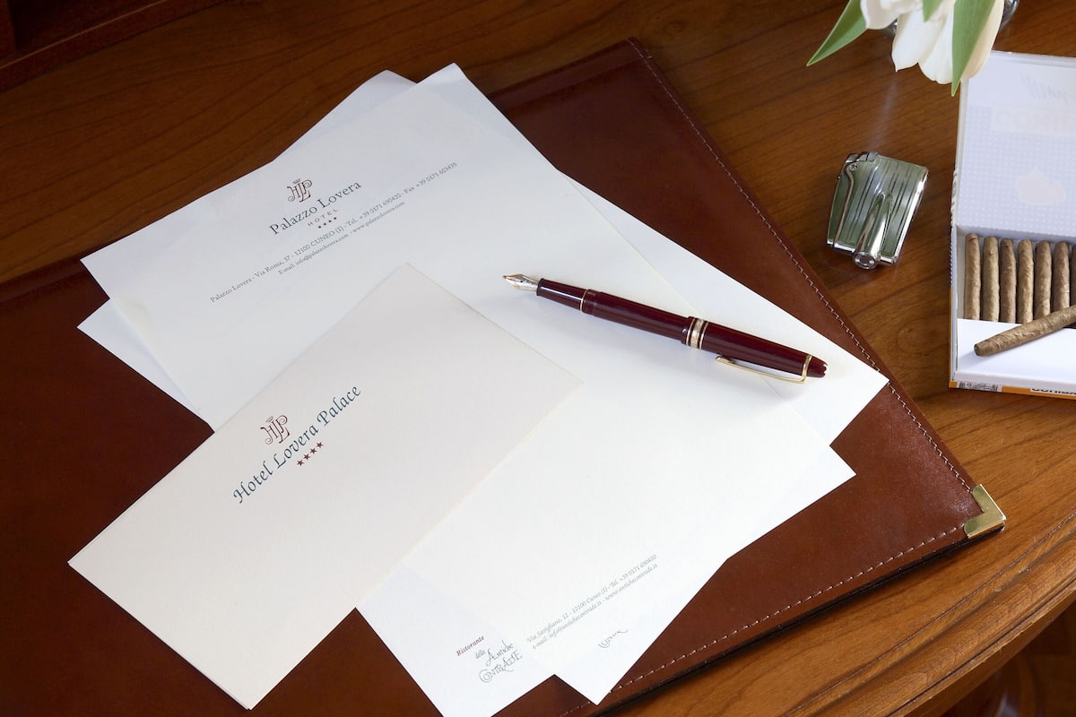 A wooden desk with a brown leather folder, two white papers, a pen, a lighter, and a box of cigarettes.