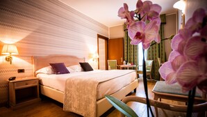 A hotel room with a large bed, bedside lamps, a wooden nightstand, a chair, and a desk with a chair. - Hotel Palazzo Lovera (Cuneo)
