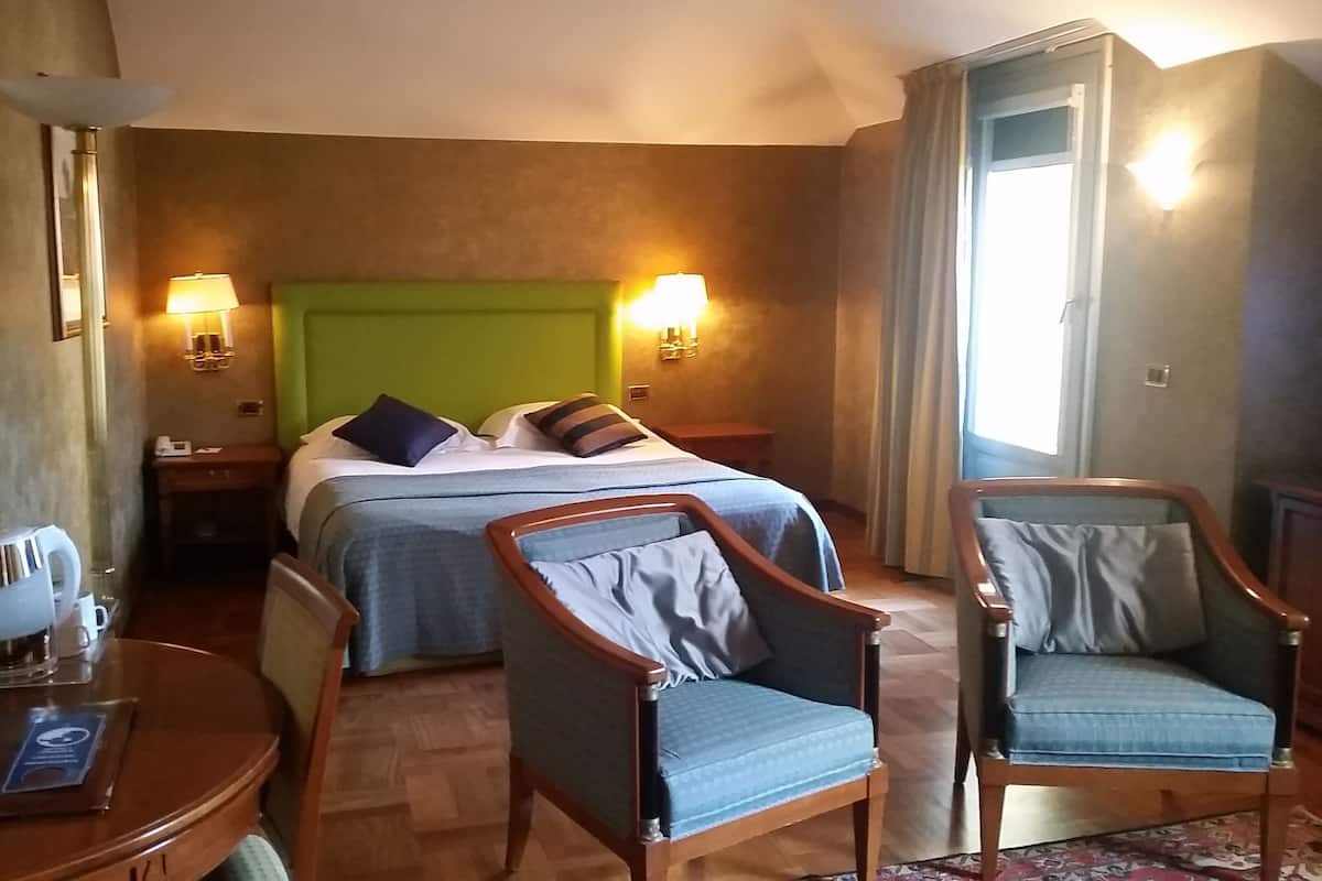 A hotel room with a bed, two chairs, a bedside table with a lamp, a mirror, and a window with curtains.