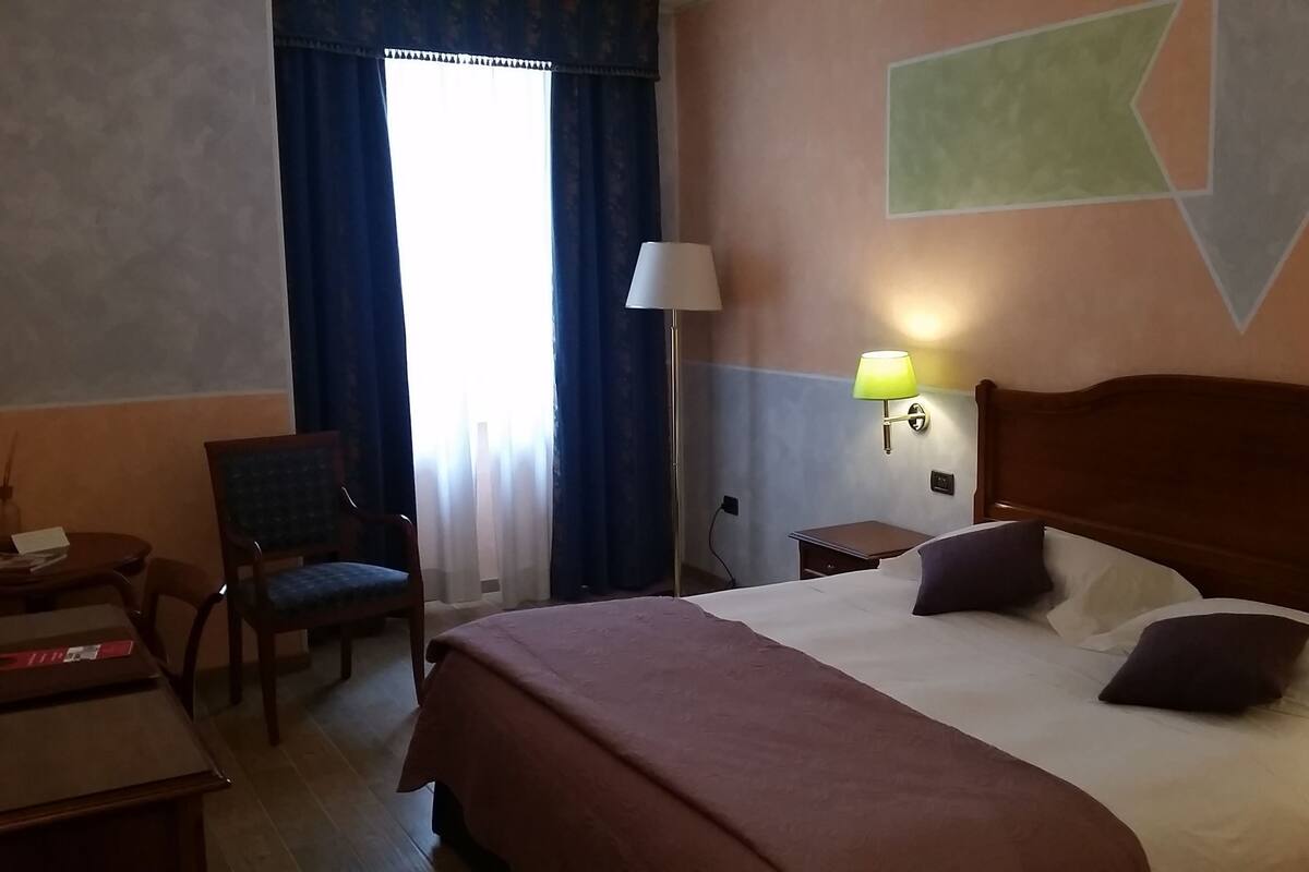 A hotel room with a bed, a chair, a desk, a lamp, and a window with curtains.