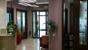 A lobby with a tiled floor, a reception desk with a clock, potted plants, and a striped chair. - Hotel Palazzo Lovera (Cuneo)