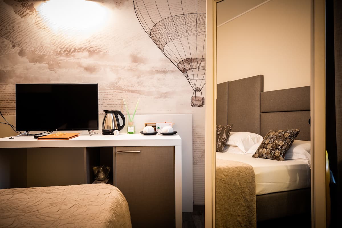 A hotel room with a bed, a desk with a computer, a kettle, and a wall art piece.