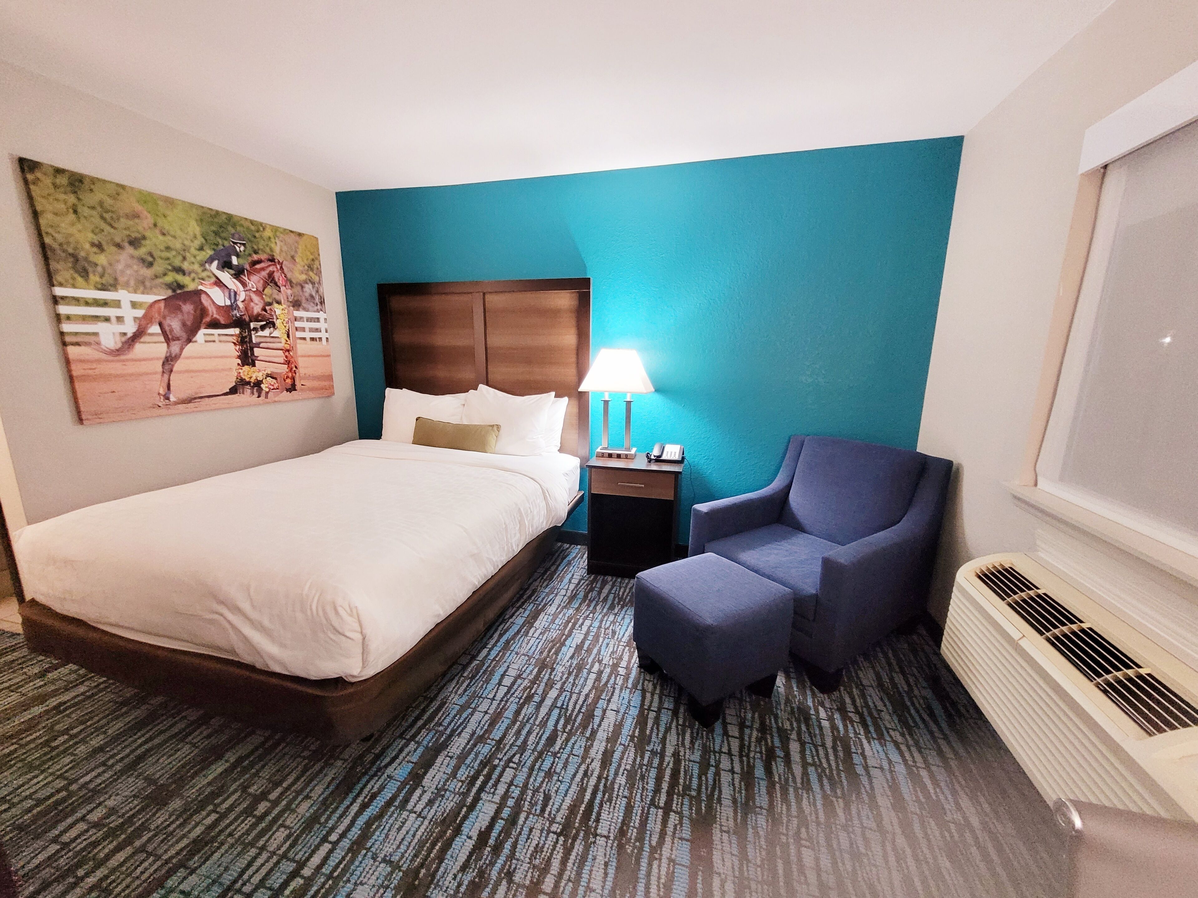 A hotel room with a large bed, a chair, a nightstand with a lamp, and a painting on the wall.