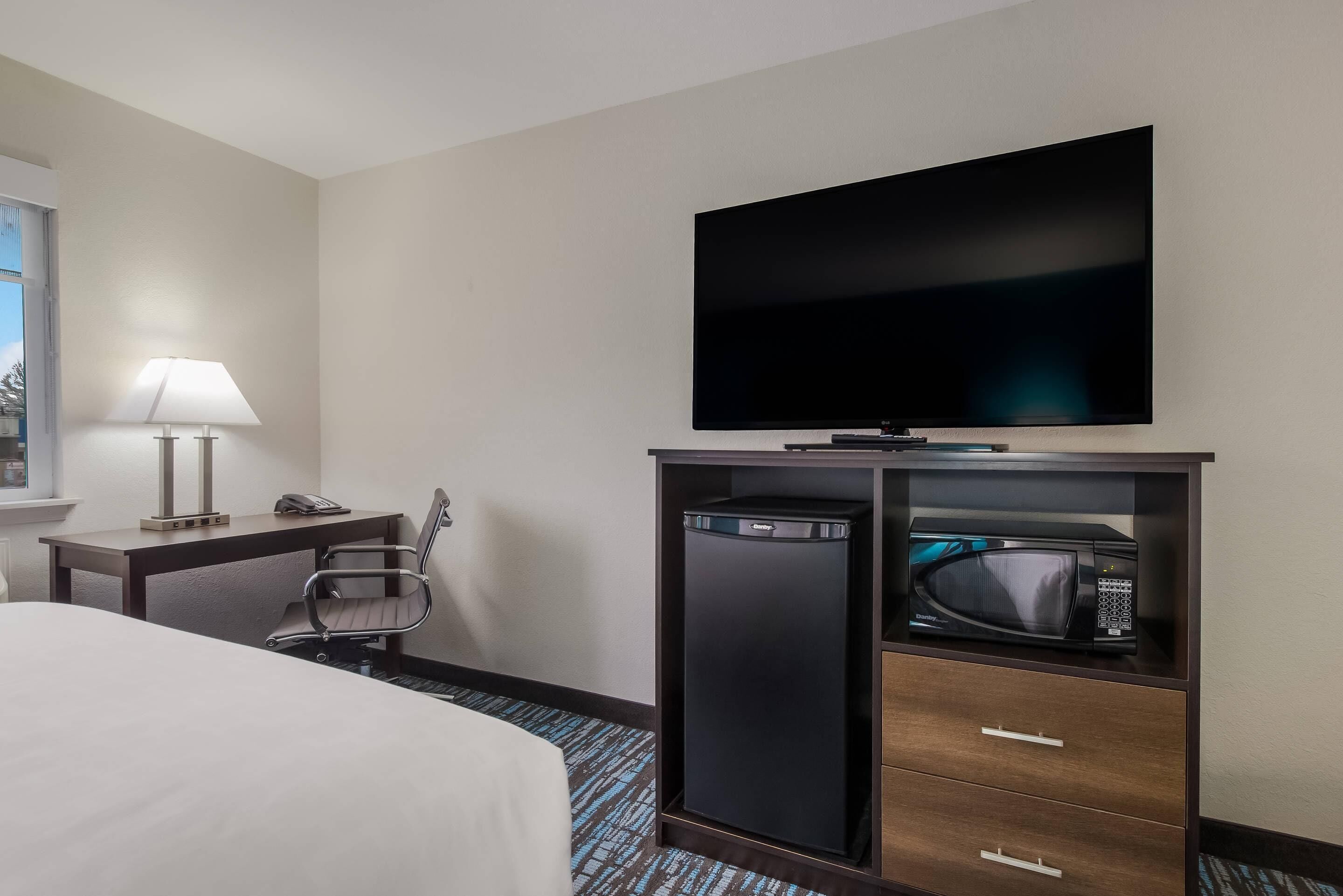 Una habitación de hotel con una cama, un escritorio con lámpara, un televisor de pantalla plana, un microondas y un mini bar.