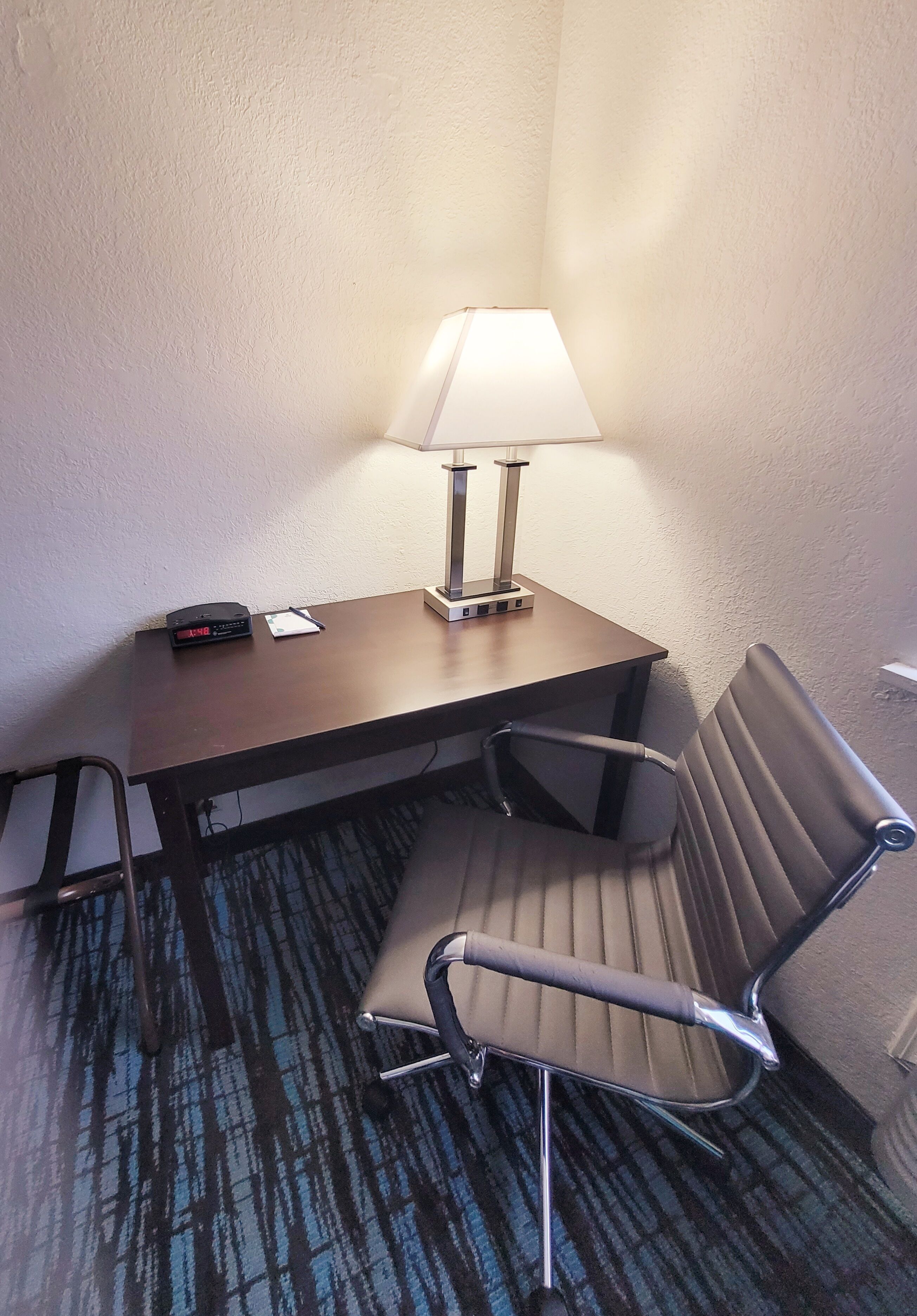 Habitación de hotel en esquina con escritorio, una silla, una lámpara y piso alfombrado.