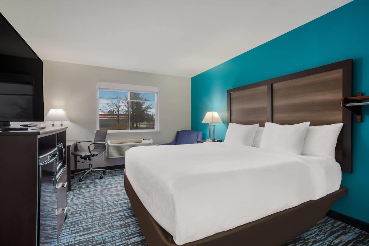 A hotel room with a large bed, a desk with a lamp, a chair, and a window with a view of trees.