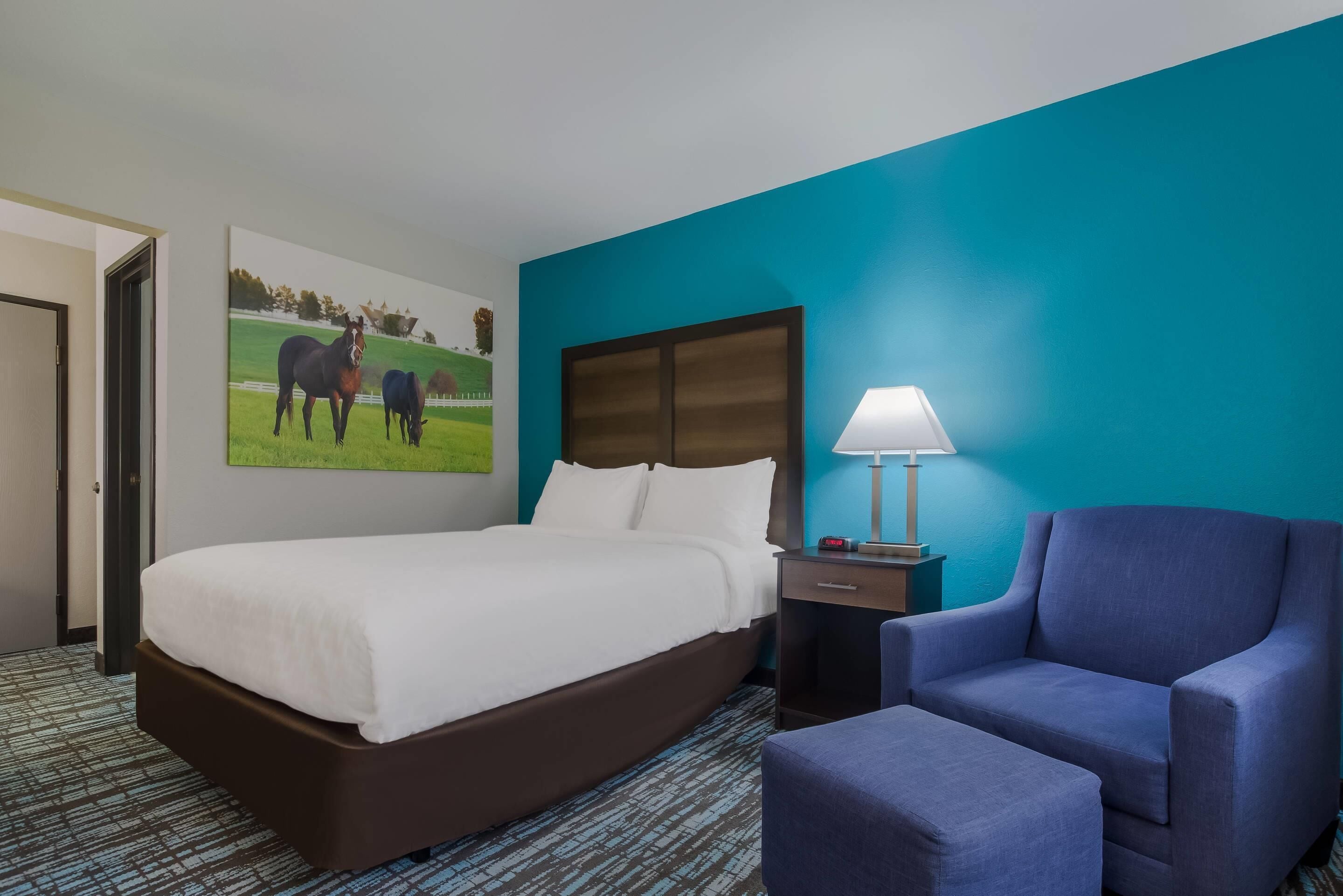 A hotel room with a bed, a chair, a nightstand, a lamp, and a painting of horses on the wall.