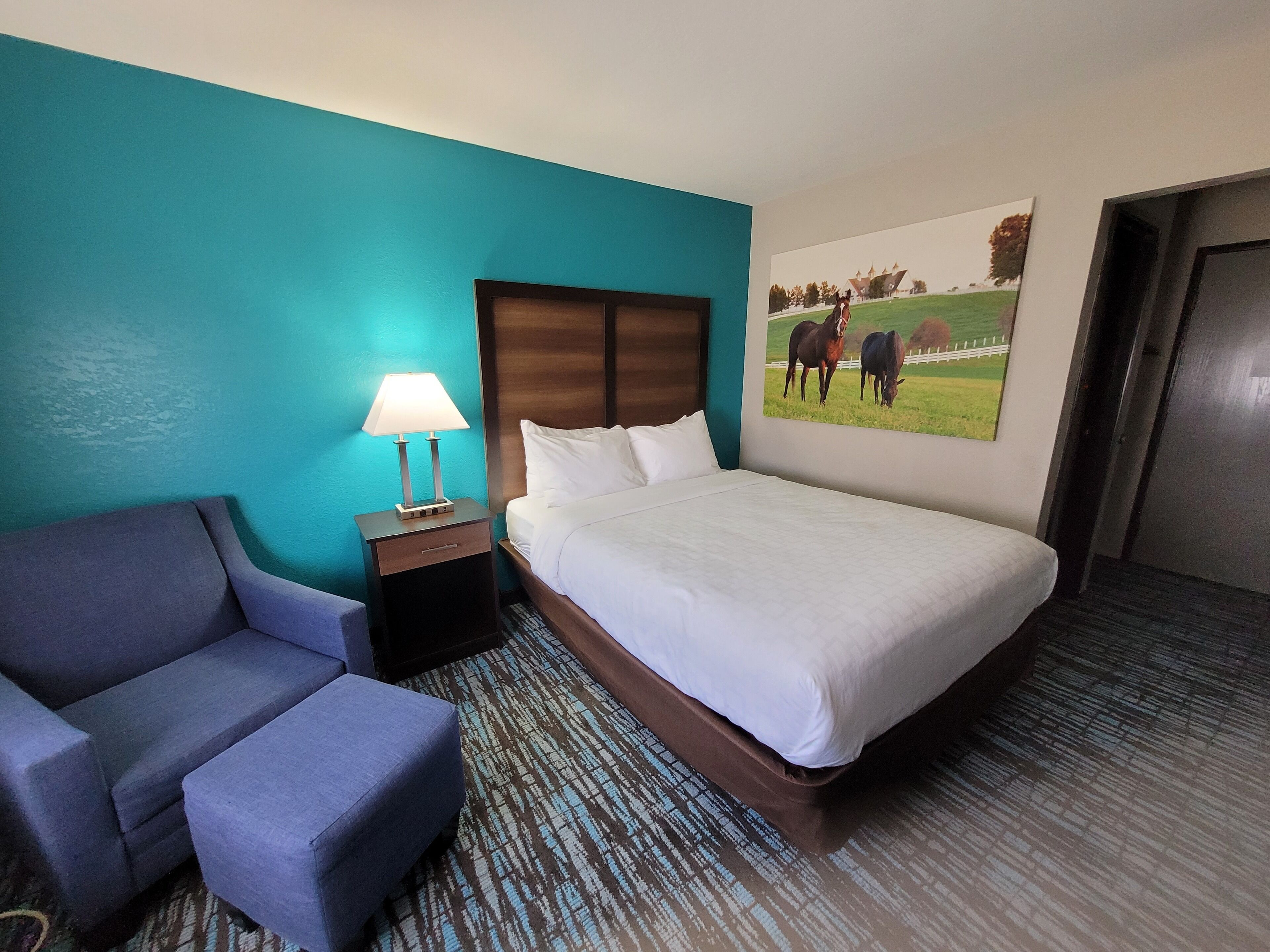 A hotel room with a bed, a chair, a nightstand with a lamp, and a painting of horses on the wall.
