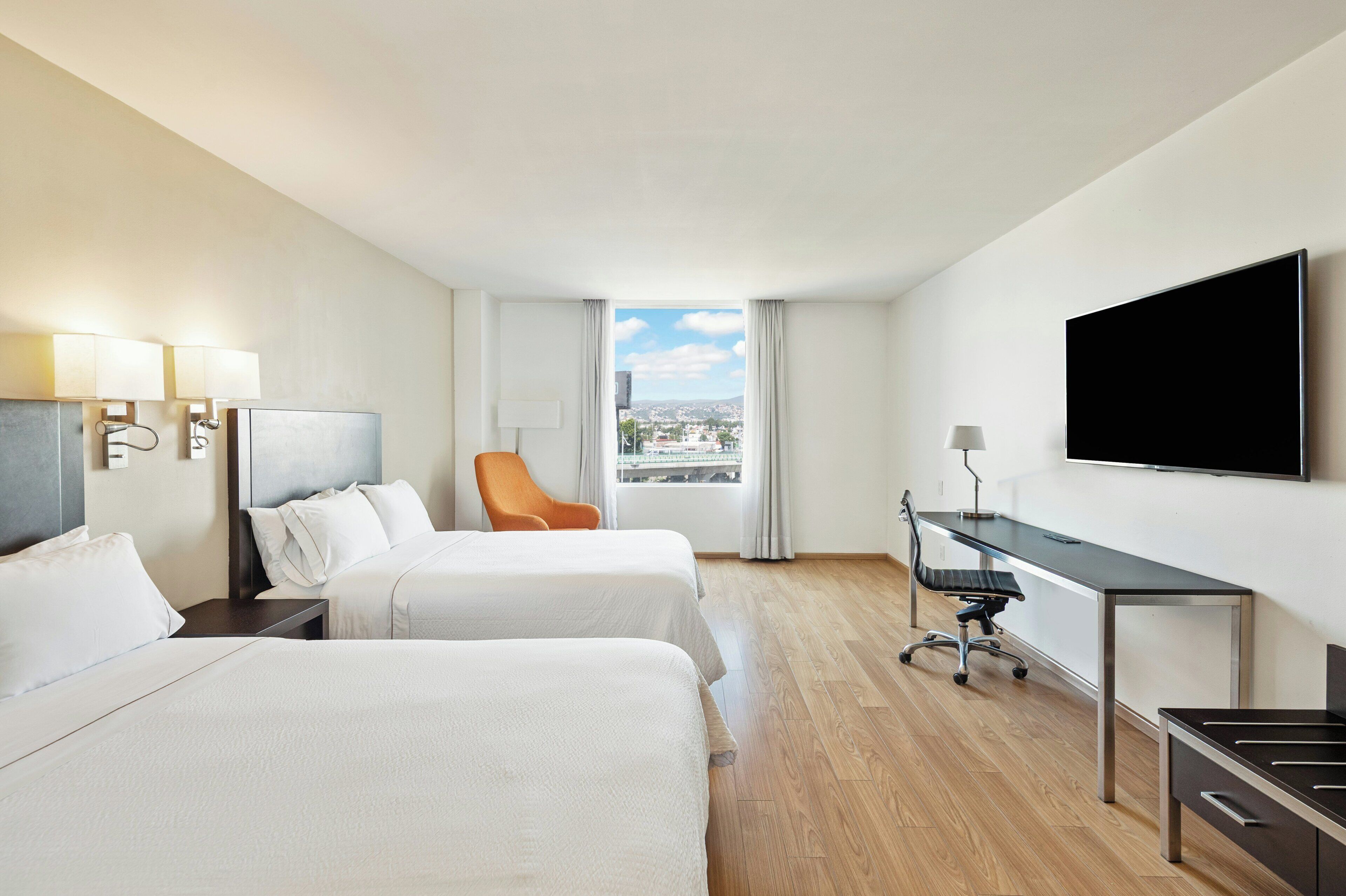 A hotel room with two beds, a desk, a TV, and a view of the city.