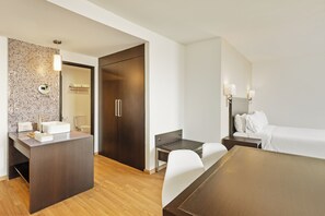 A modern hotel room with a wooden floor, a large bed, a small desk with a mirror, and a bathroom with a tiled wall and a wooden door.