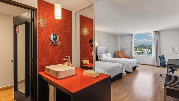 A hotel room with a red-tiled wall, a large window with a city view, a bed with two pillows, a desk with a chair, and a bathroom with a sink and mirror. - Fiesta Inn Express Naucalpan (Naucalpan)