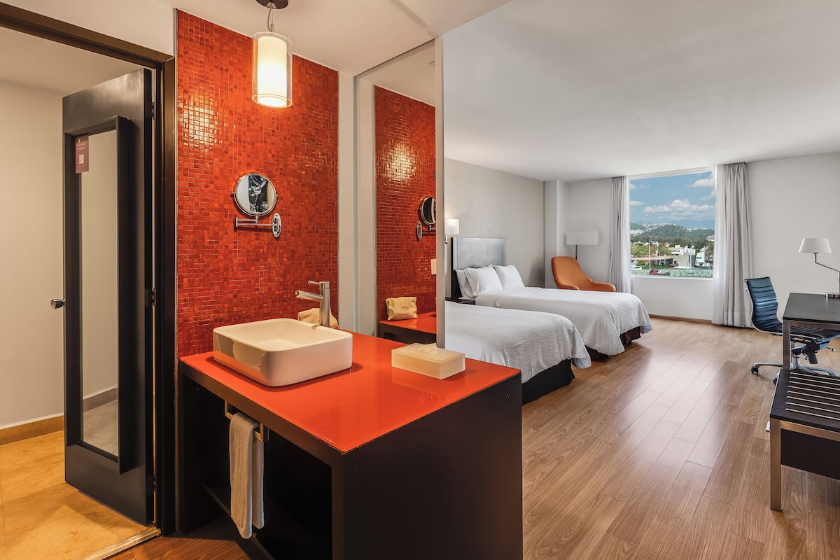 A hotel room with a red-tiled wall, a large window with a city view, a bed with two pillows, a desk with a chair, and a bathroom with a sink and mirror.