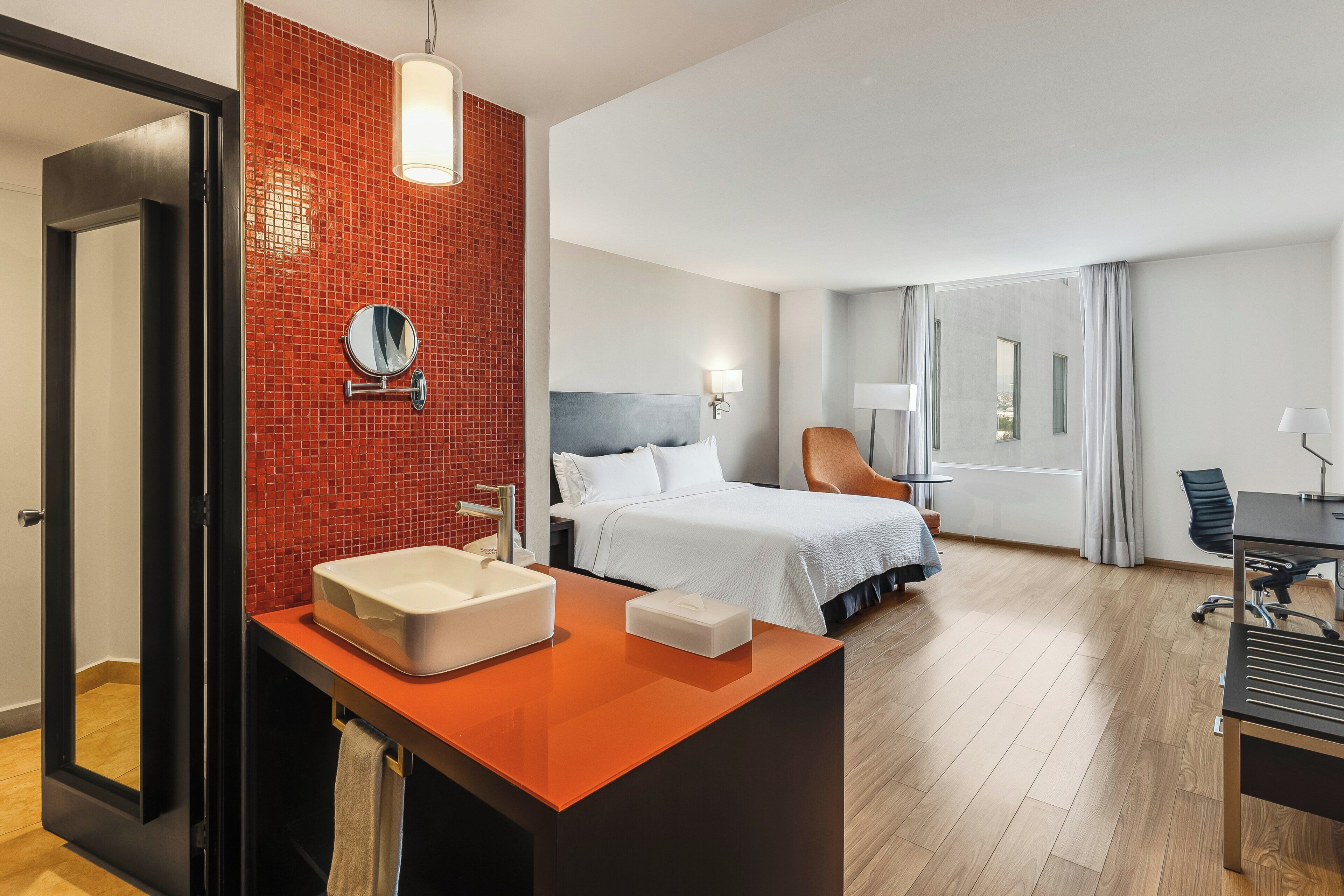 Une chambre d’hôtel comprenant un lit, un bureau, une chaise, une salle de bain avec un lavabo et un miroir, et un mur recouvert de carreaux rouges.