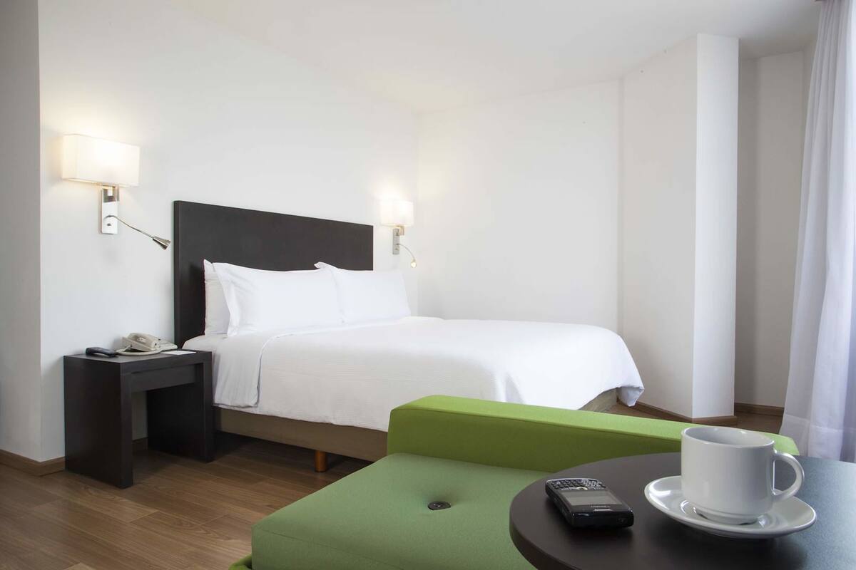 A hotel room with a bed, bedside table with a phone, a green ottoman, a table with a cup and remote control.