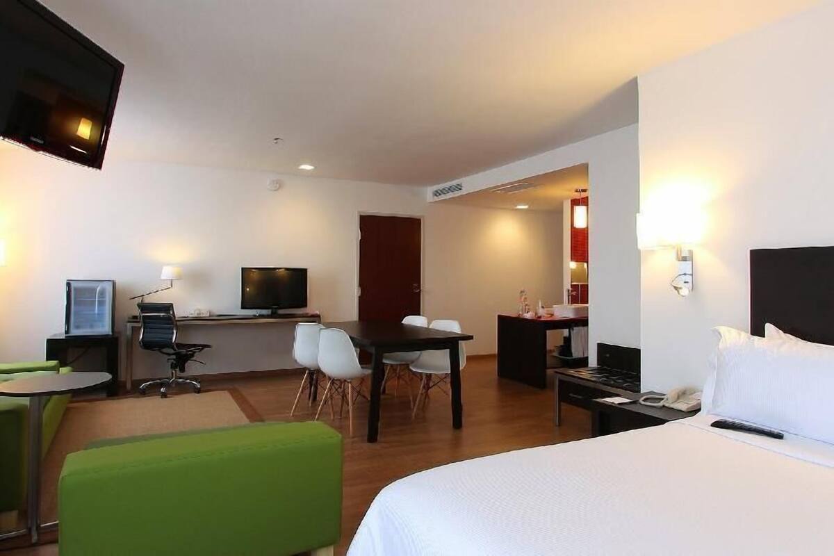 Een hotelkamer met een bed, een bureau met computer, een televisie, een eettafel met stoelen en een groene bank.