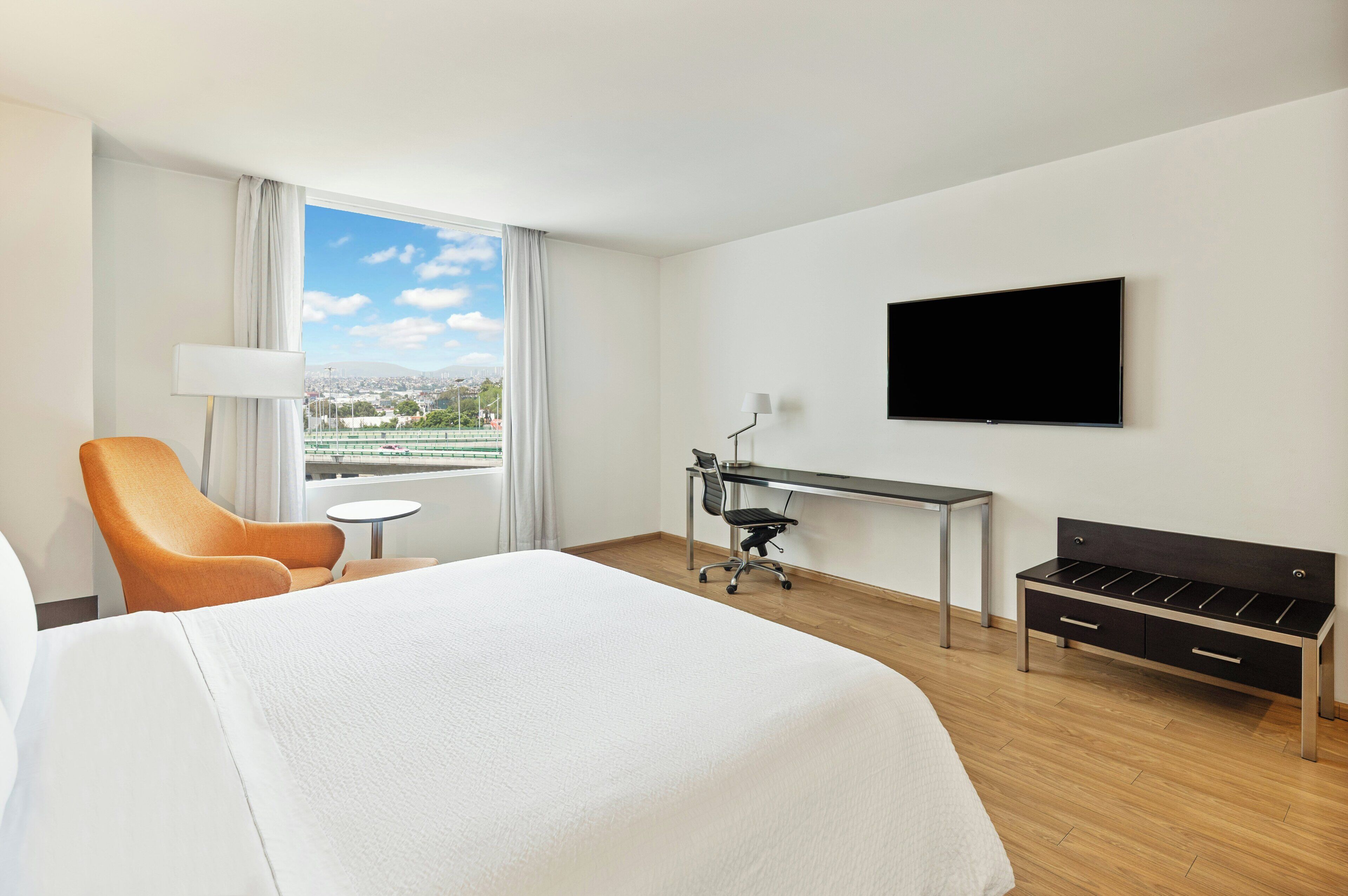 A hotel room with a bed, a desk, a chair, a TV, and a view of a cityscape.
