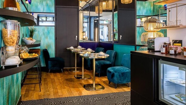 Un interior de restaurante moderno con paredes verde azulado, suelo de madera y una combinación de mesas redondas y cuadradas.