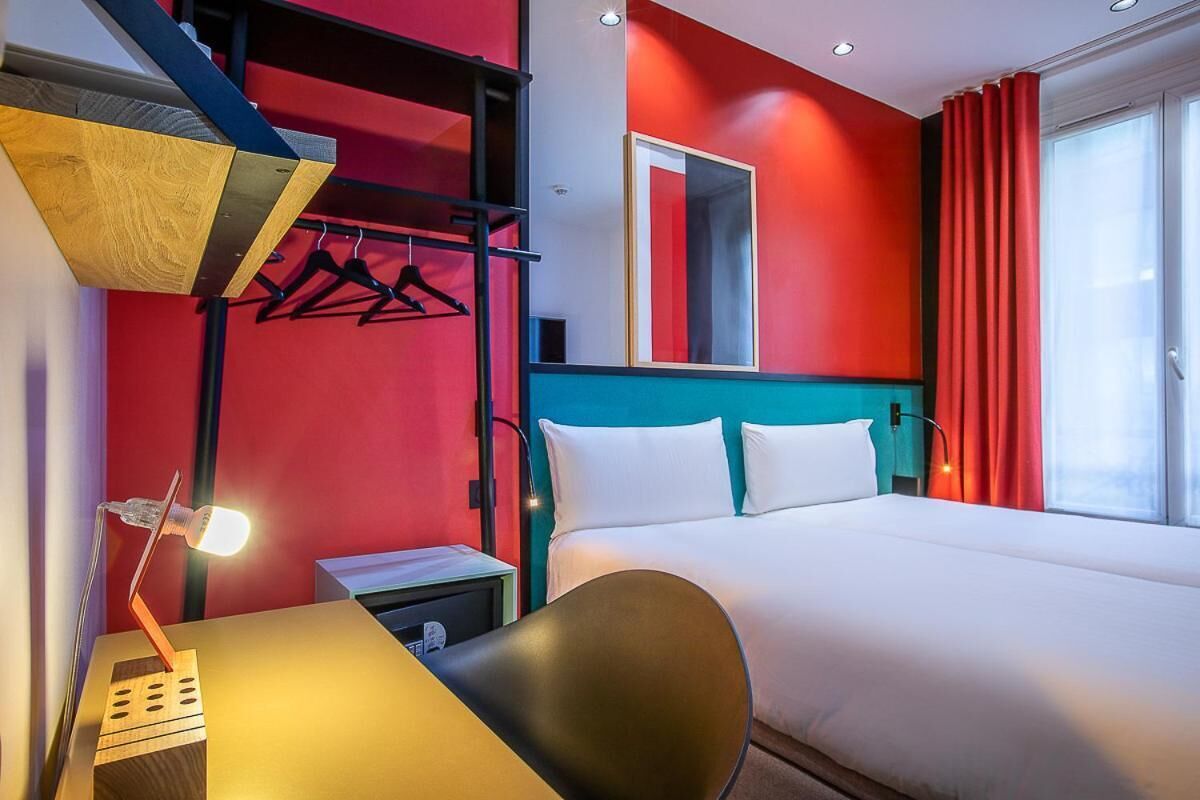 A hotel room with a bed, a desk with a lamp, a chair, a coat rack, and a window with curtains.