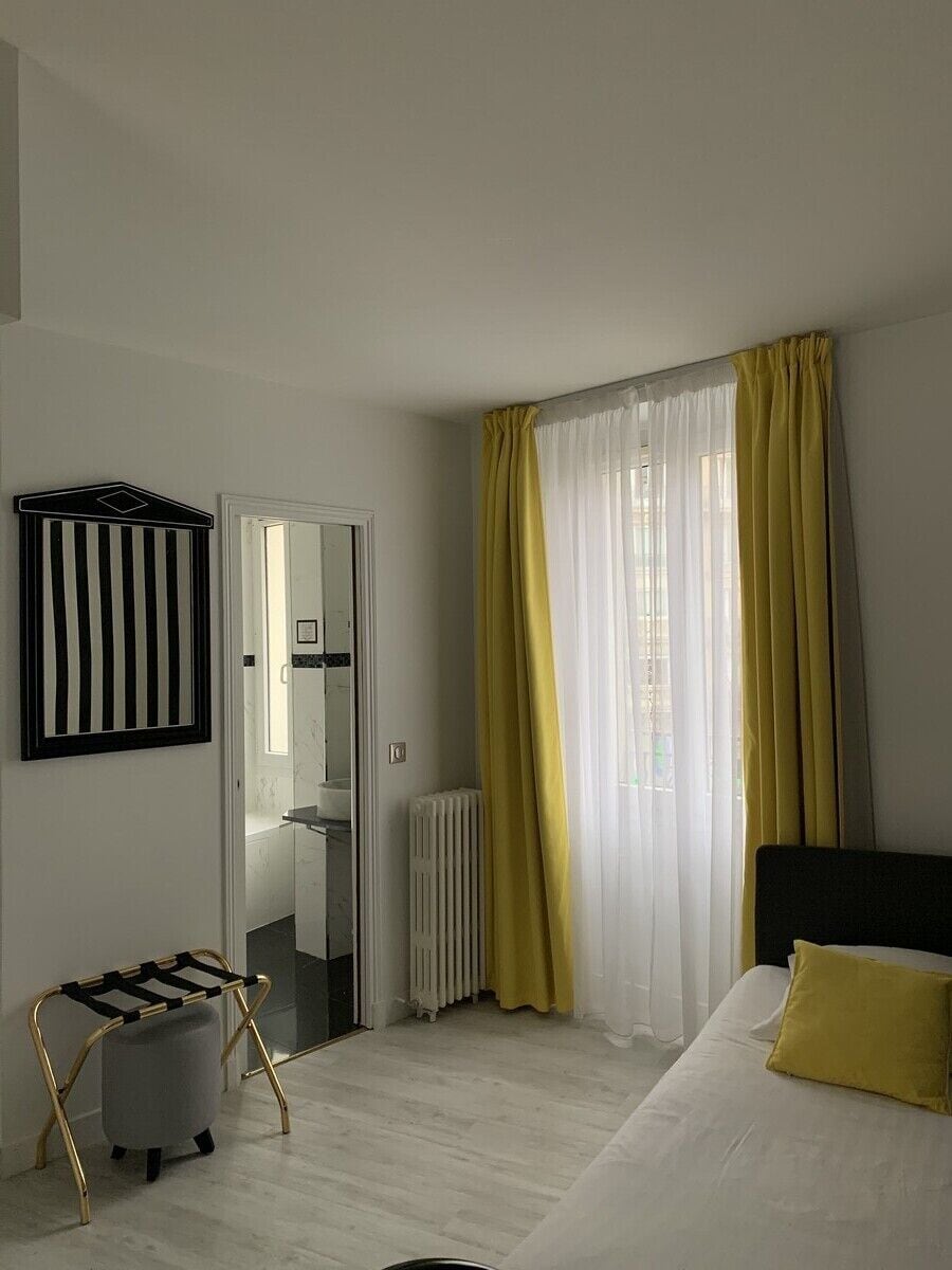 A hotel room with a bed, a radiator, a mirror, and a window with yellow curtains.