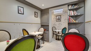 A dining area with tables and chairs, a bookshelf, and framed pictures on the walls. - Hotel Riviera Elysées (Paris)