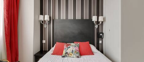 Une chambre d’hôtel avec un lit, des rideaux rouges, un mur à rayures et deux lampes fixées au mur.
