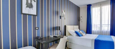 Una habitación de hotel con paredes a rayas, un escritorio con lámpara, una cama con manta azul y un cuadro en la pared.
