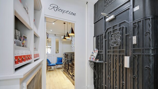 A reception area with a sign, a blue chair, and a display of items on shelves. - Hotel Riviera Elysées (Paris)