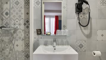 Un baño con lavamanos blanco, cabina de ducha de cristal, secador de pelo montado en la pared y azulejos estampados.