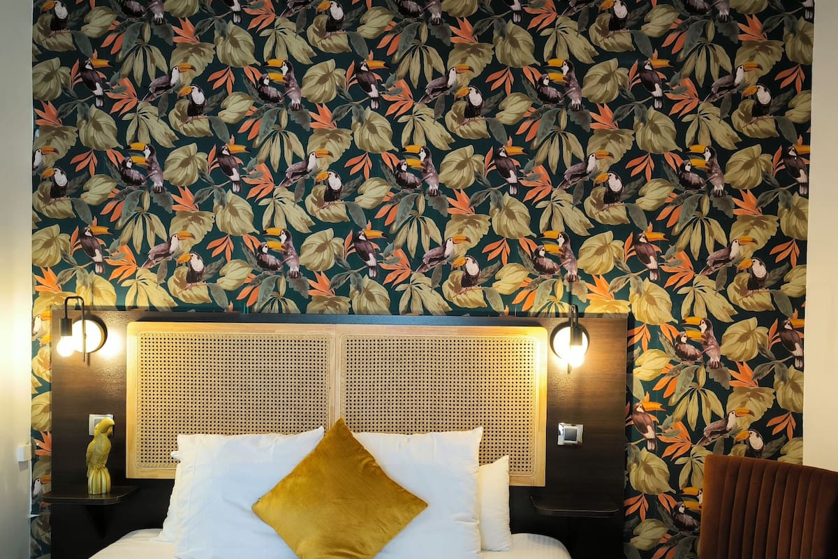 Una habitación de hotel con una cama, cabecero con diseño tejido, pared con motivos de hojas tropicales y pájaros, y una silla marrón.