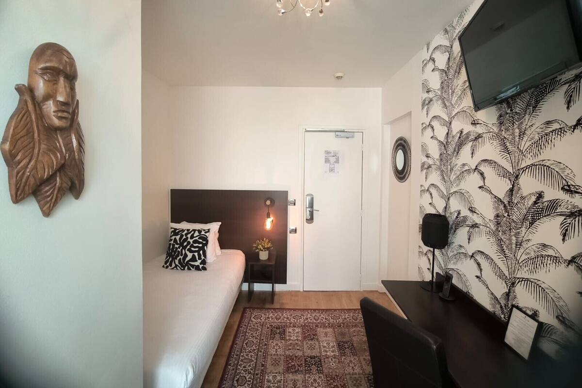 Habitación de hotel con cama, mesita de noche, escritorio, candelabro, televisor montado en la pared y un papel tapiz estampado.