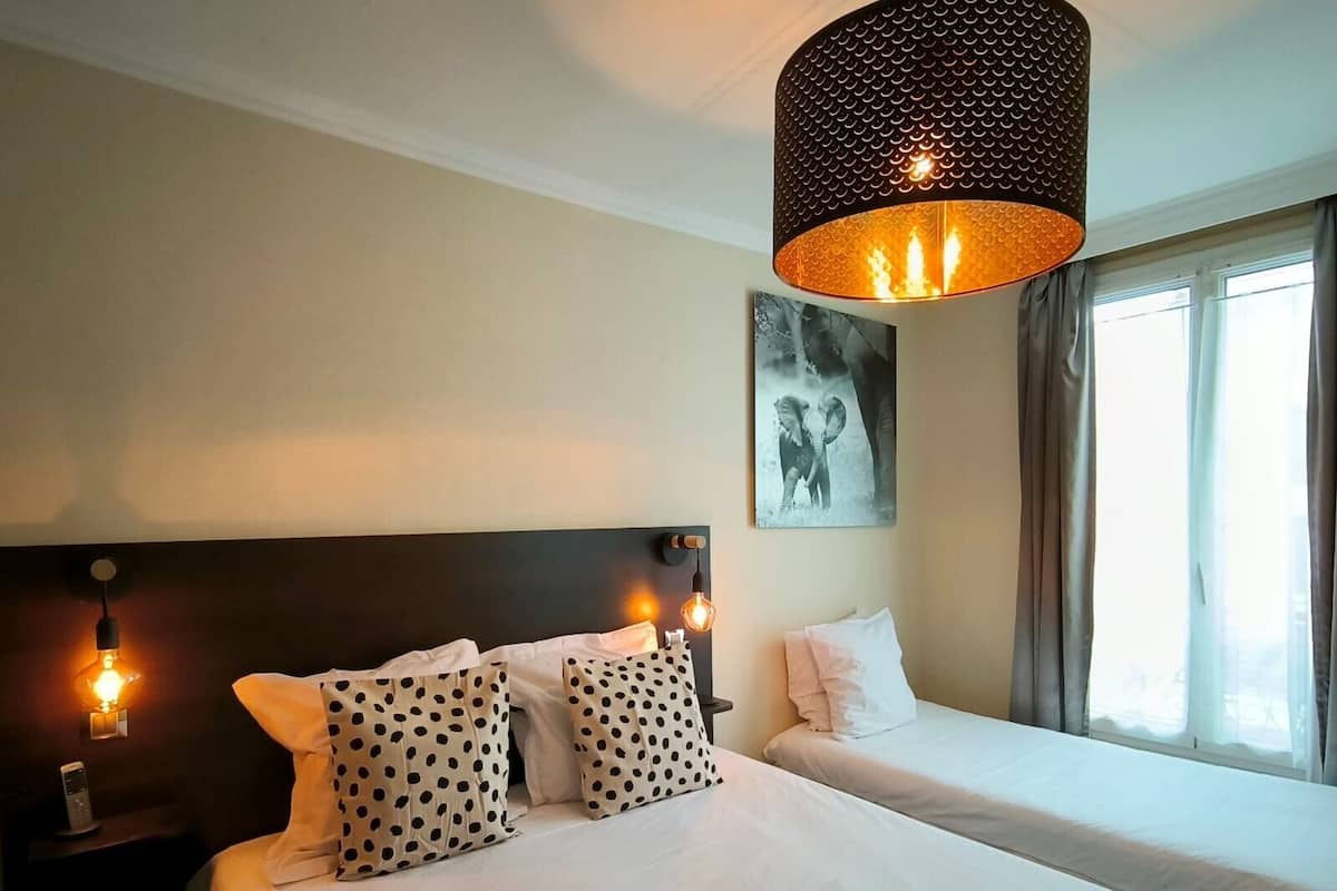 Habitación de hotel con una cama grande, dos mesitas de noche, una lámpara colgante y un ventanal con cortinas.
