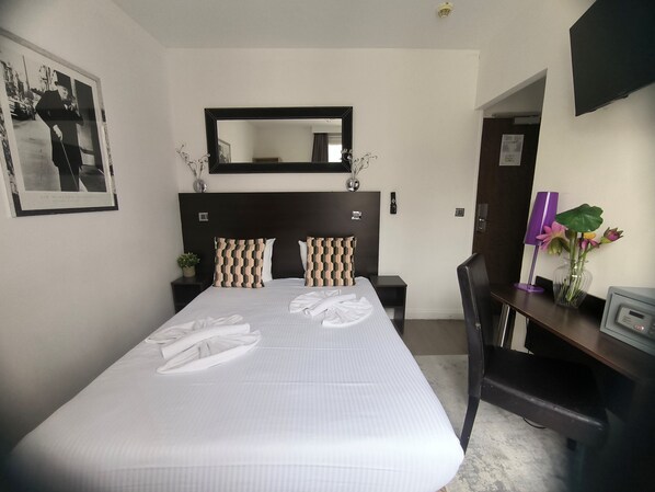 A hotel room with a large bed, a desk, a chair, a mirror, and a wall-mounted TV.