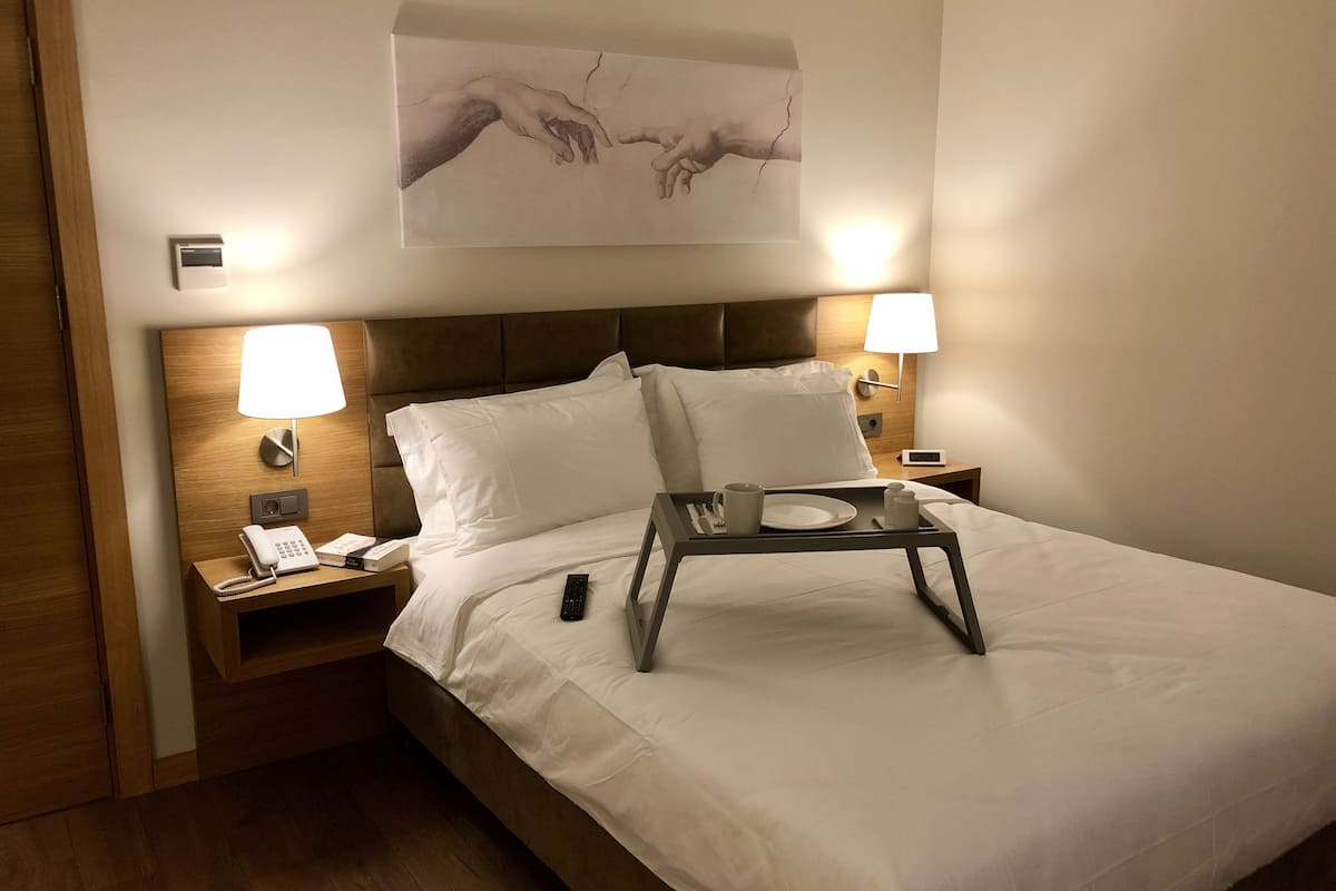 A neatly made bed with white linens, two bedside lamps, a bedside phone, a tray with a plate, a cup, and a remote control.