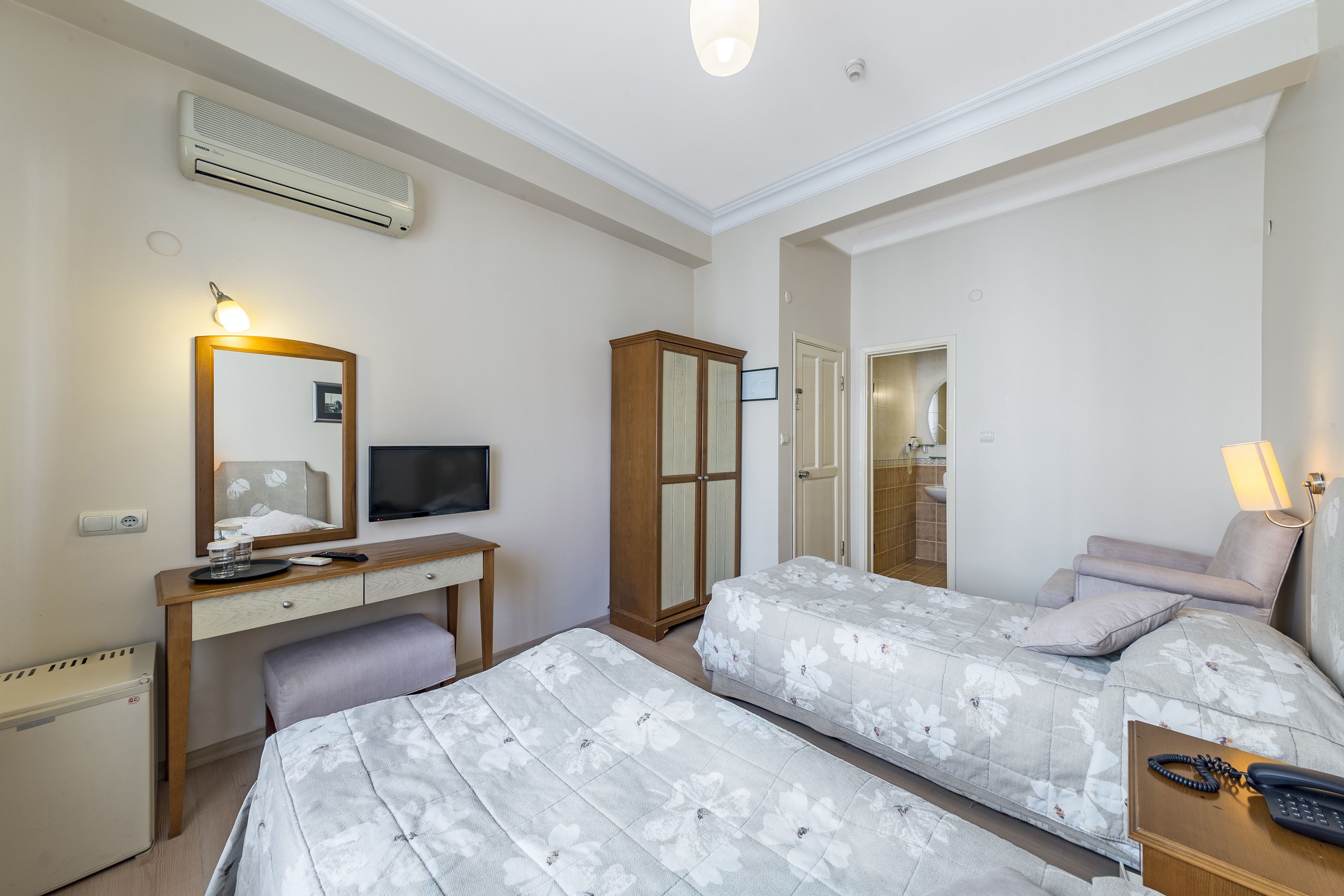 Une chambre d’hôtel avec deux lits, un bureau avec un ordinateur, une armoire et une salle de bain visible à travers une porte ouverte.