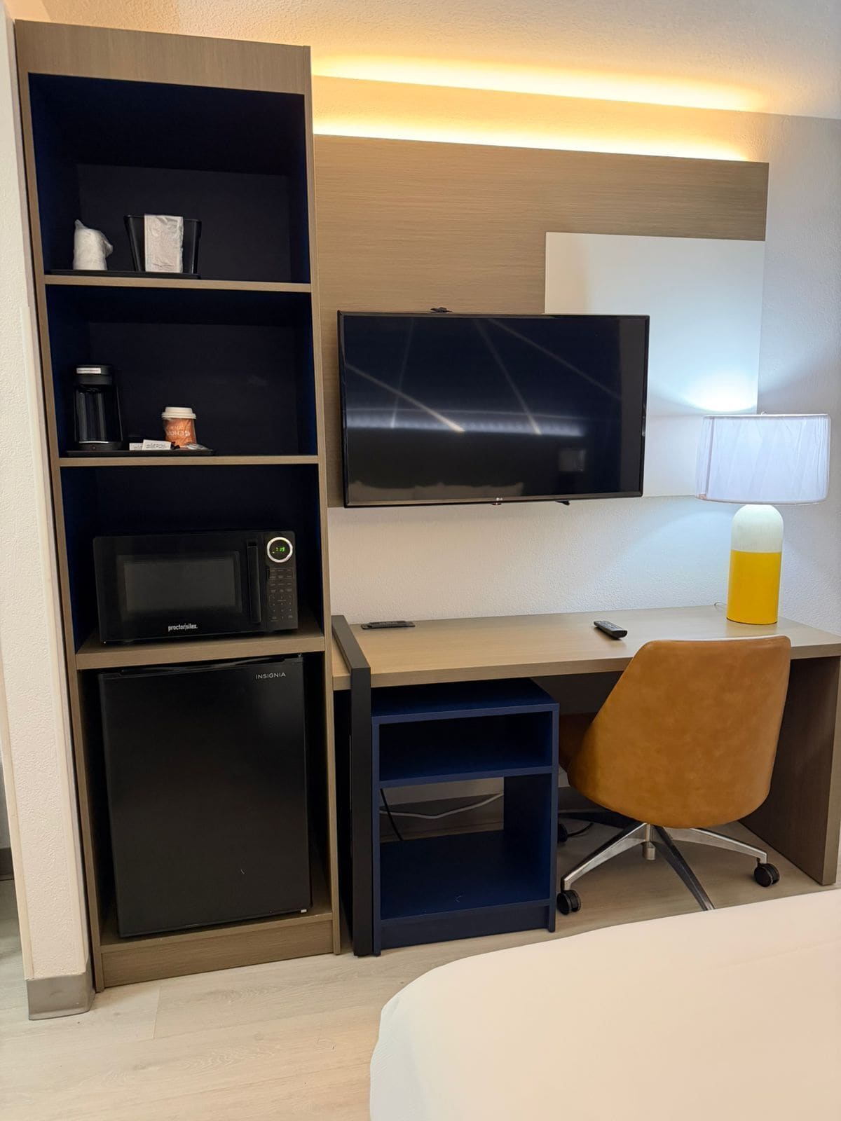 A hotel room with a flat-screen TV, a coffee maker, a microwave, a mini fridge, a desk, a chair, and a bedside table.
