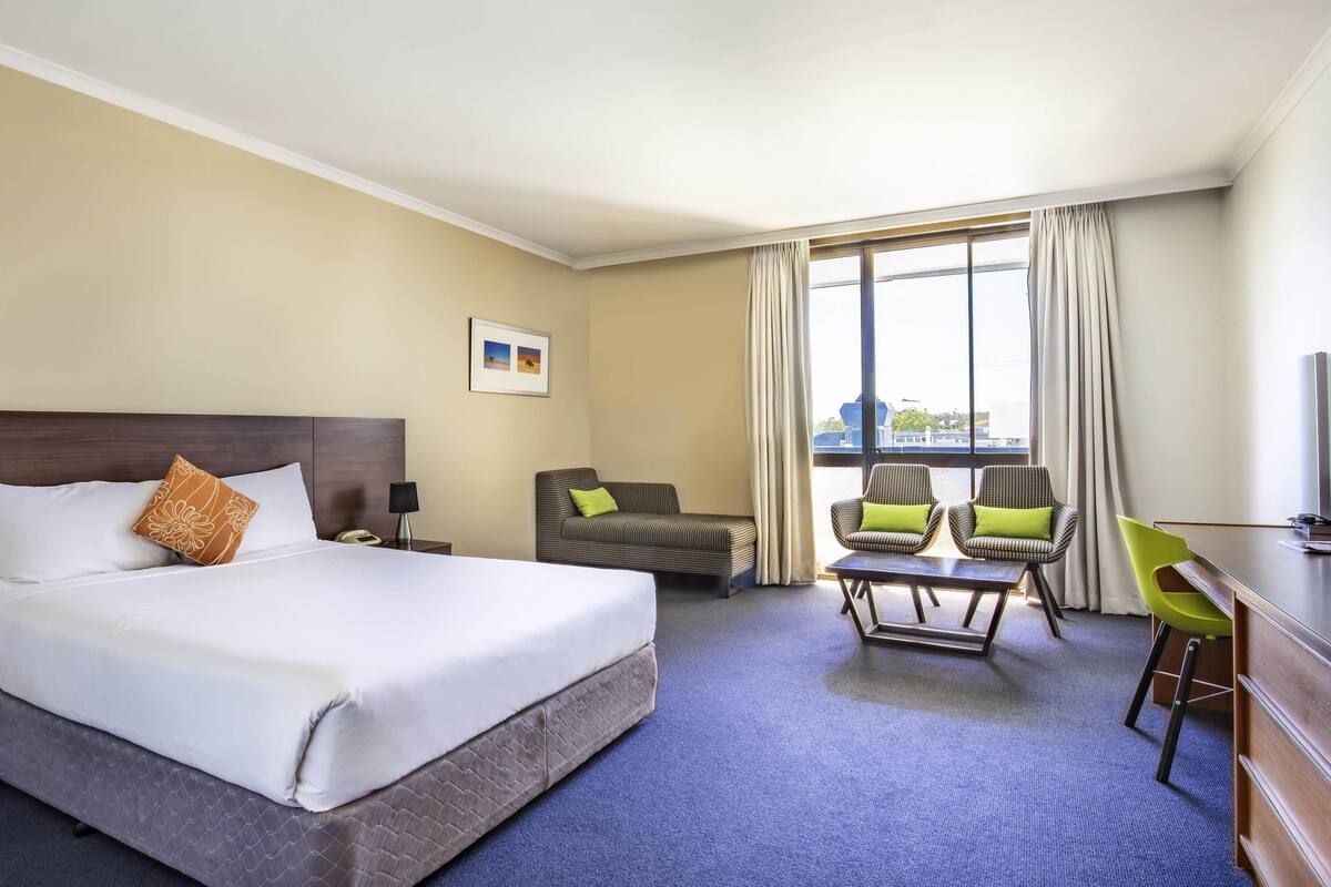Habitación de hotel con una cama grande, un escritorio, dos sillas, una mesita y una ventana con cortinas.