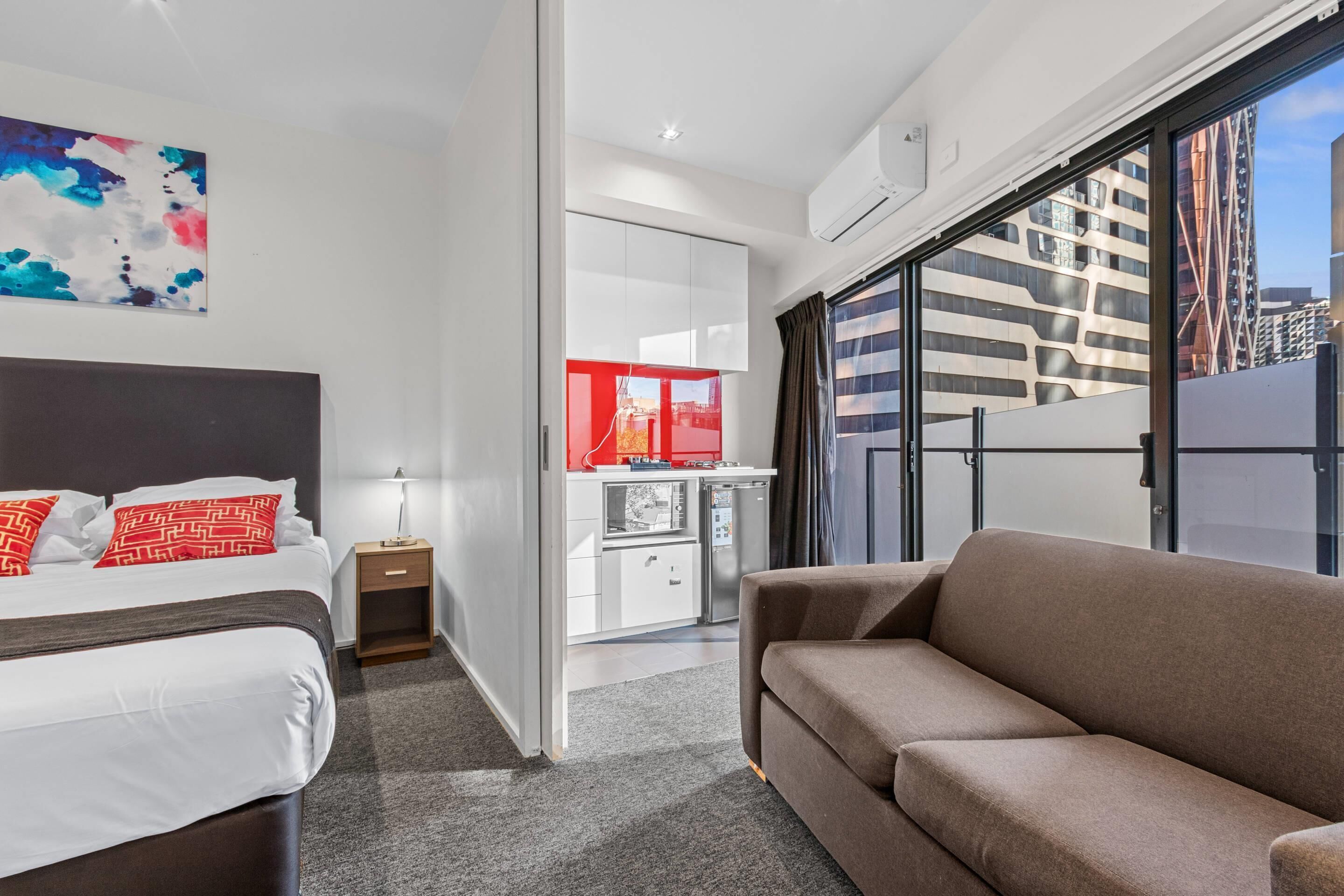 Una habitación de hotel moderna con cama, un sofá marrón, kitchenette y un ventanal con vistas a edificios.
