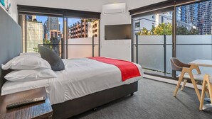 A modern hotel room with a large bed, a TV mounted on the wall, a balcony with a view of the city, and a small table with a chair. - Comfort Apartments on Elizabeth (Melbourne)