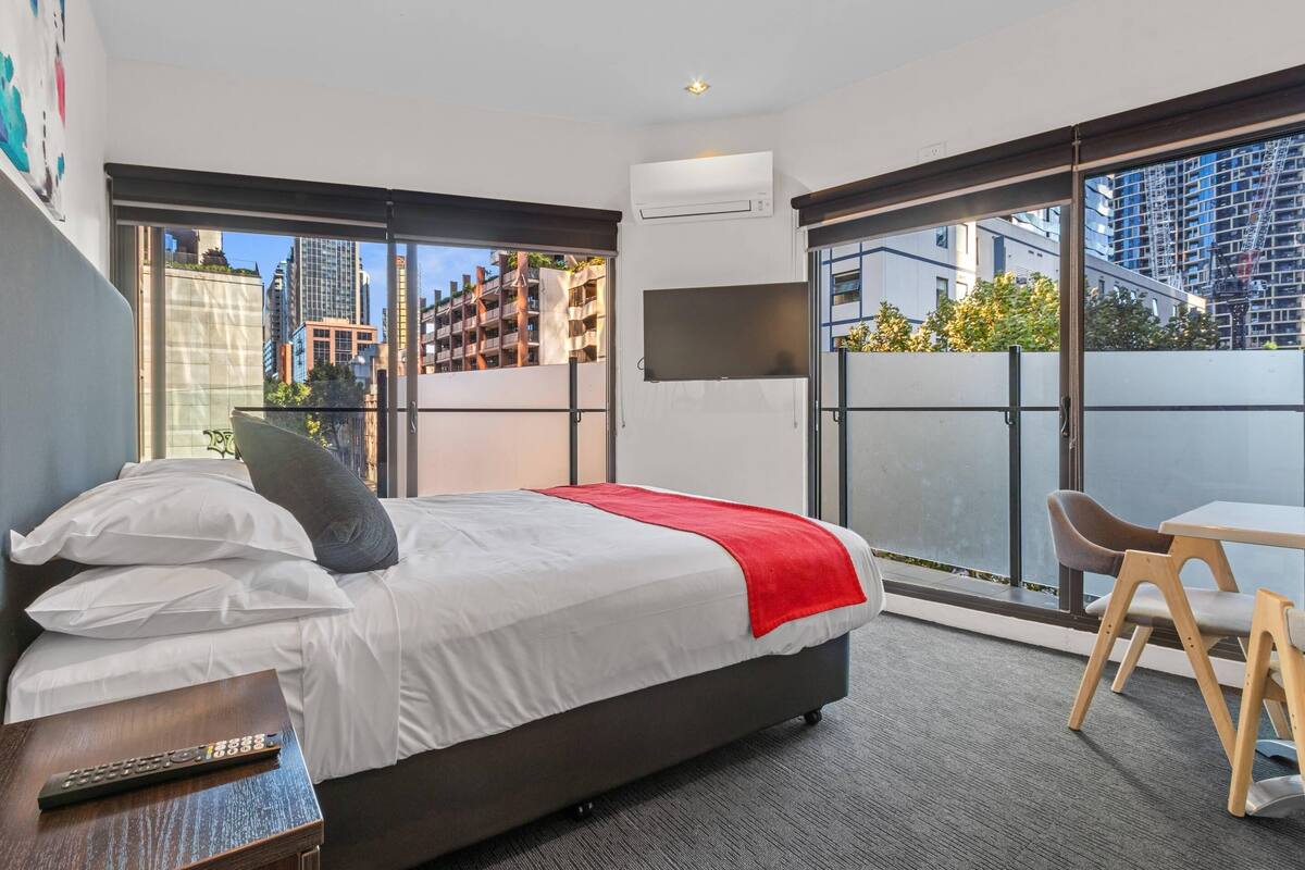 Una habitación de hotel moderna con una cama grande, un televisor montado en la pared, un balcón con vistas a la ciudad y una pequeña mesa con una silla.