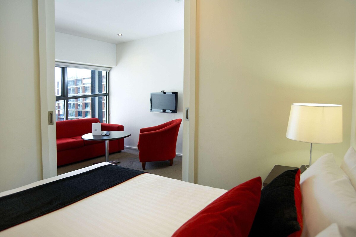 A hotel room with a bed, a TV, a red armchair, a small table, and a lamp.