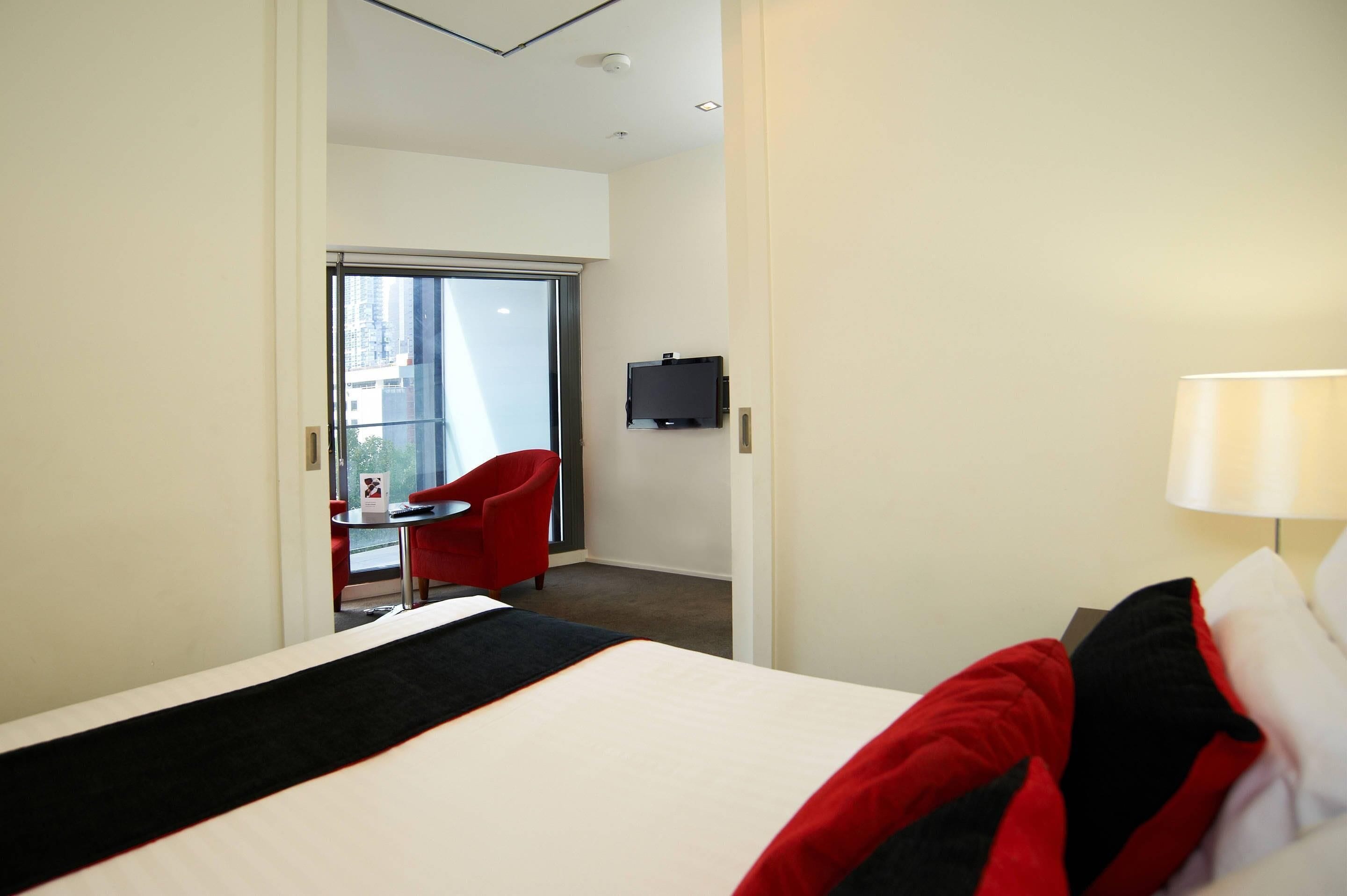 A hotel room with a bed, a red chair, a small table, a TV, and a balcony with a red chair.