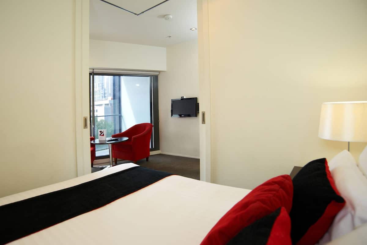 A hotel room with a bed, a red chair, a small table, a TV, and a balcony with a red chair.
