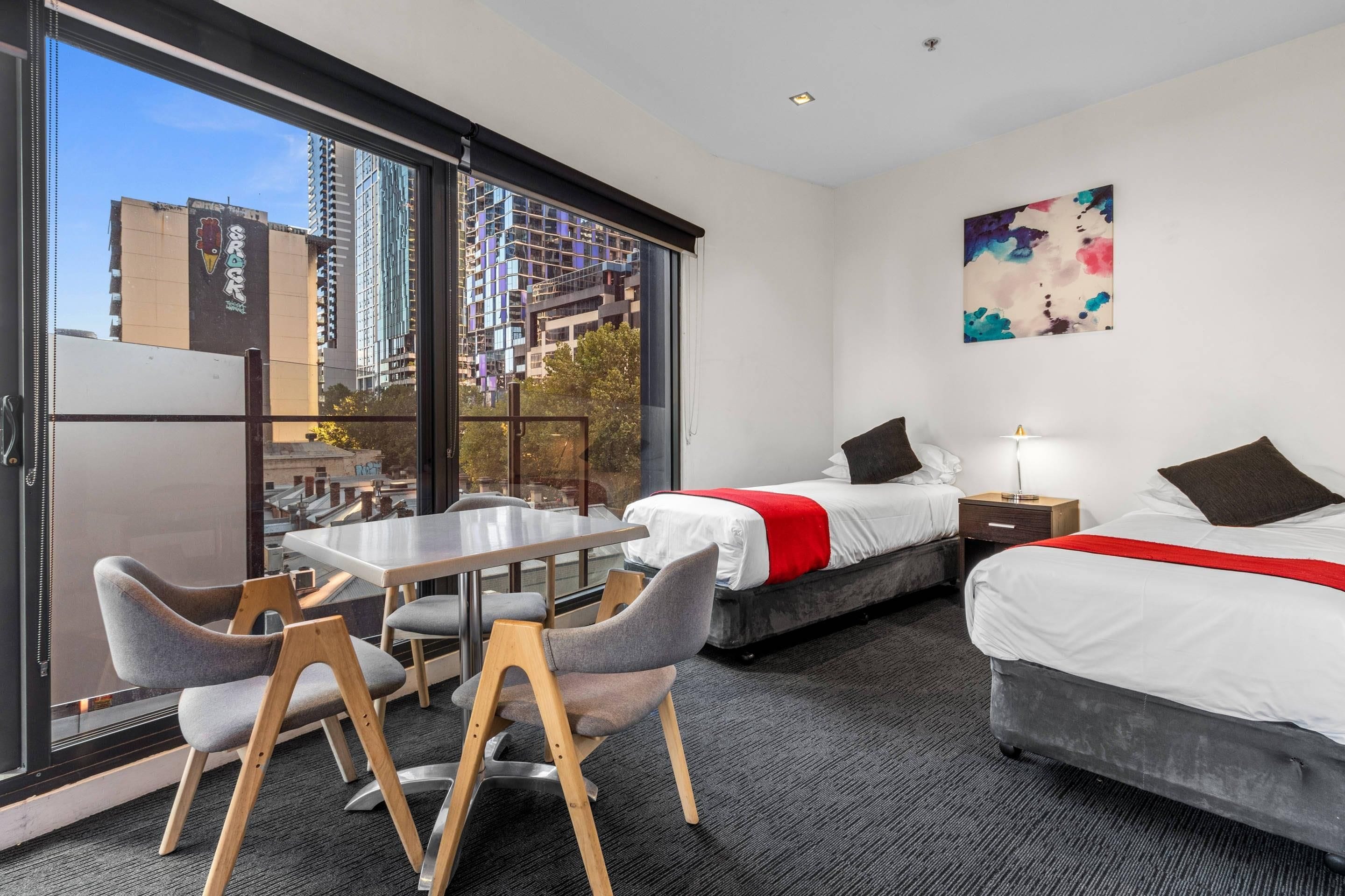Een hotelkamer met twee bedden, een eettafel met stoelen, een groot raam met uitzicht op de stad en een schilderij aan de muur.