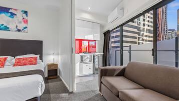 A modern hotel room with a bed, a sofa, a small kitchenette, and a large window with a city view.