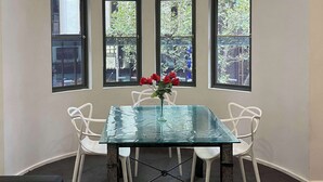 A glass-top table with four white chairs, a vase with red flowers, and three windows with a view of greenery. - Comfort Apartments on Elizabeth (Melbourne)