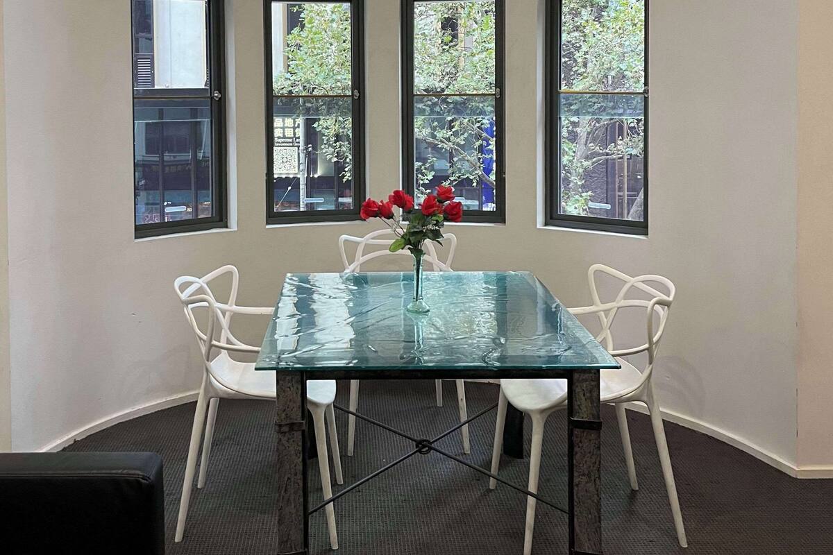 Una mesa con superficie de vidrio, cuatro sillas blancas, un florero con flores rojas y tres ventanas que dan a vistas de vegetación.