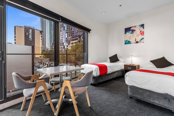 A hotel room with two beds, a dining table with chairs, a large window with a city view, and a painting on the wall. - Comfort Apartments on Elizabeth (Melbourne)
