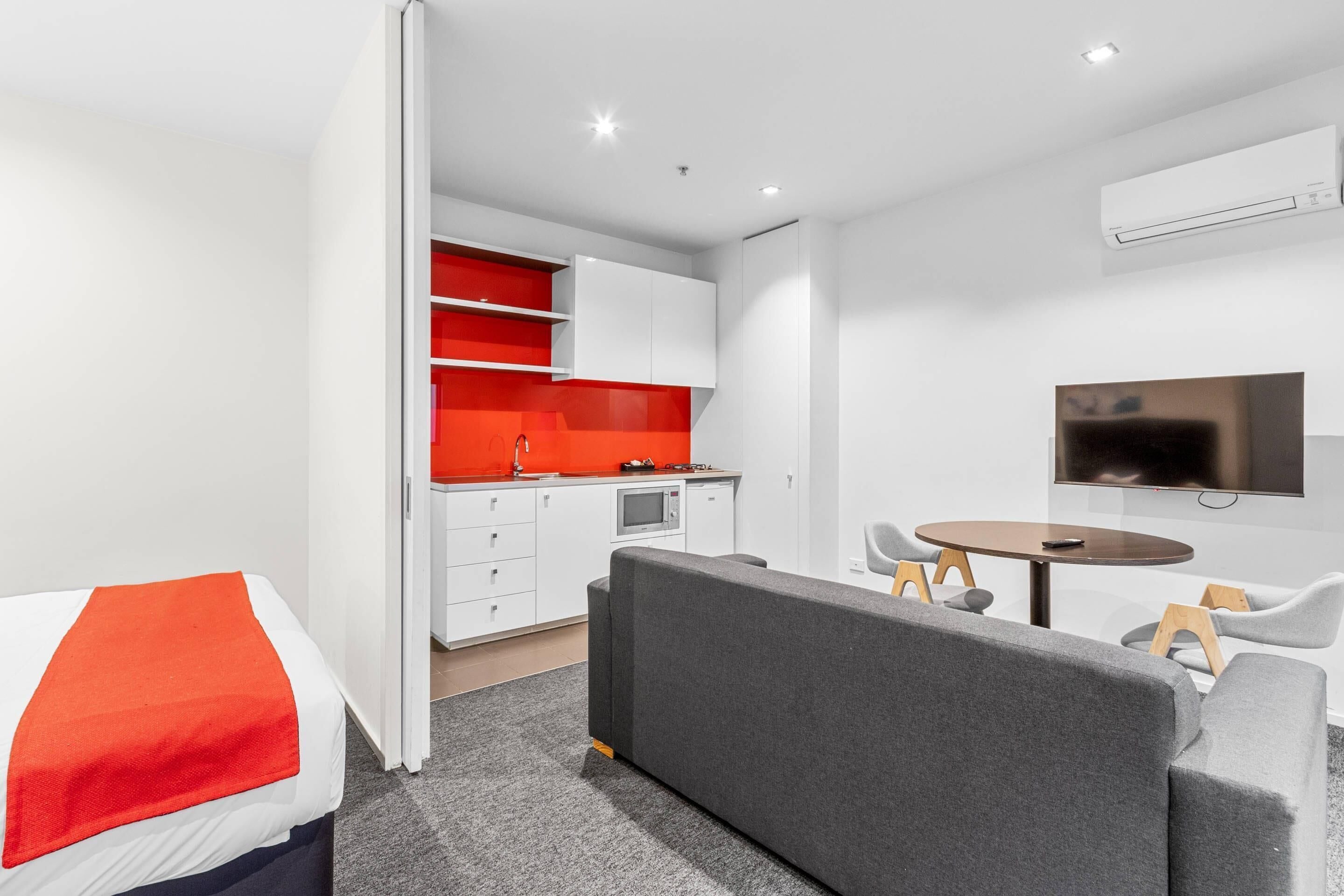 A modern hotel room with a bed, grey sofa, dining table, and kitchenette.